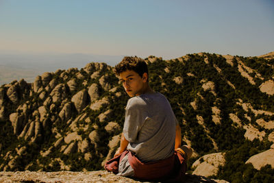 Boy sitting on rock against clear sky