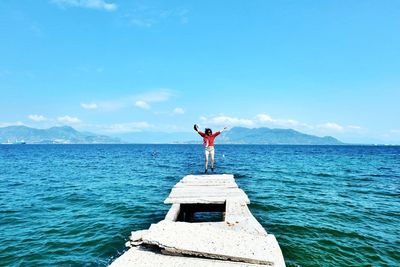 Woman jumping on old pier on sea against blue sky