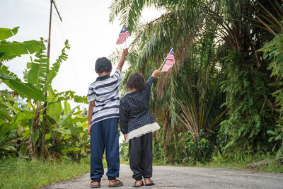 Rear view siblings holding malaysian flag while standing on road amidst plants