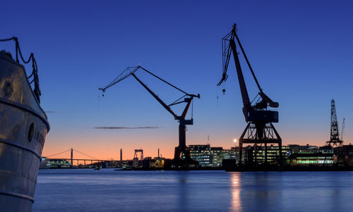 Cranes at harbor against blue sky during sunset