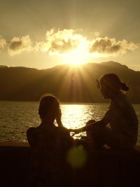 Rear view of couple sitting on shore against sunset sky