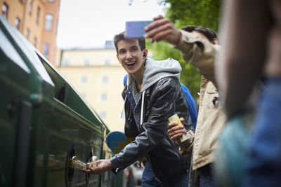 Man taking selfie with teenage friend throwing waste in garbage bin at recycling station