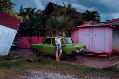 Young lady in casual wear laughing while standing near green old car among rural houses and tropical plants in cuba