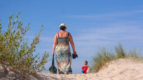 Rear view of mother and son walking at beach against blue sky