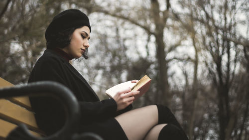 Woman reading book while sitting on bench against trees