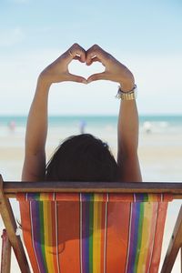 Woman gesturing heart shape while sitting on deck chair at beach against sky