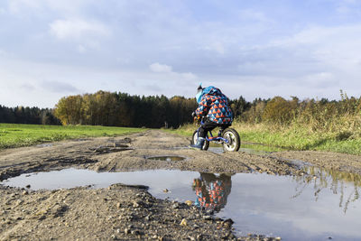 Child riding bicycle on puddle against cloudy sky