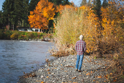 Teen boy in jeans, plaid shirt, and beanie walks along river in fall.