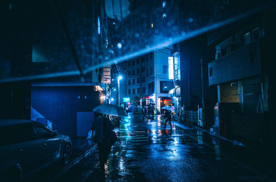 People walking on illuminated street amidst buildings during rainy season