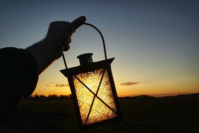 Cropped hand of man holding lantern against sky during sunset