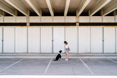 Full body side view of young female in casual outfit standing with big dog near metal wall of urban building