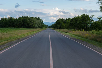 Empty road amidst trees on field against sky