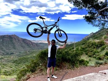 Full length of man lifting bicycle while standing on mountain against sky