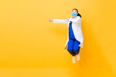 Full length of woman wearing mask jumping against yellow background