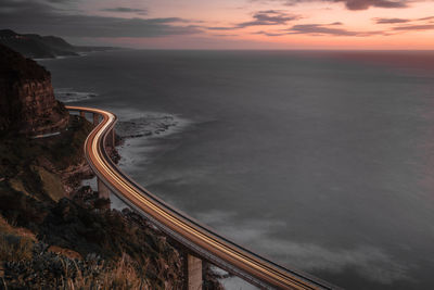 Aerial view of light trail on bridge by sea against cloudy during sunset
