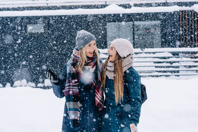 Two young women in warm winter clothes stand under the snowfall and laugh merrily.