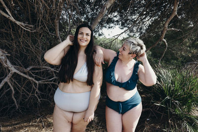 Cheerful curvy female friends in swimwear standing on beach near trees in summer