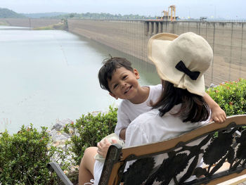 Portrait of smiling boy with mother sitting on bench against river