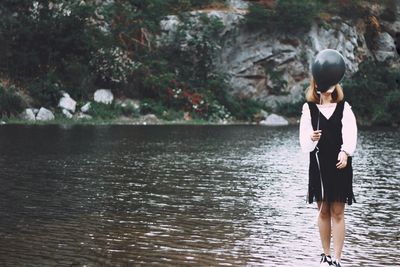 Full length of woman standing by lake and holding black balloon