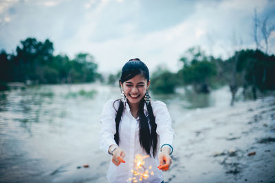 Cheerful woman holding lit sparklers at lakeshore