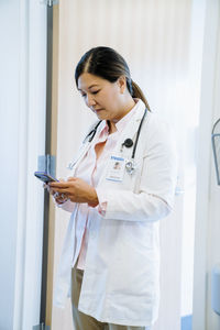 Female doctor using smart phone while standing against door in hospital