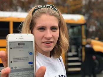 Portrait of young woman showing mobile phone while standing against school bus