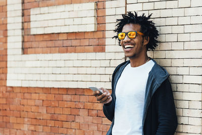 Smiling man holding smart phone while standing against brick wall