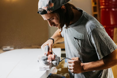 Male shaper using electric planer and polishing surface of surfboard in workshop