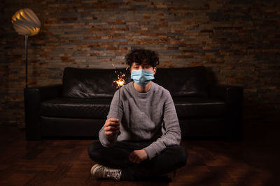 Portrait of man wearing mask holding sparkler sitting at home