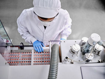 From above female factory worker in uniform operating conveyor with pills in pharmaceutical manufacturing laboratory