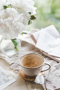 Stylish ceramic cup with  coffee and foam at window with curtains and glass vase peonies, cozy home