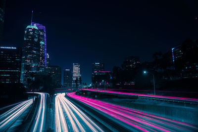 Light trails on street amidst illuminated buildings against sky at night