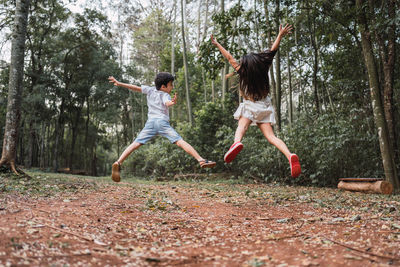 Back view of anonymous joyful boy with sister jumping with outstretched arms over terrain with trees