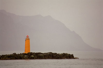 Lighthouse on rocks against mountain during foggy weather