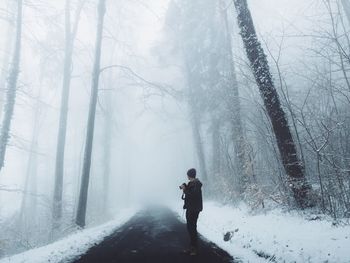 Man with camera standing on road amidst trees in forest during winter