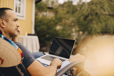 Thoughtful man using laptop while sitting at holiday villa during summer