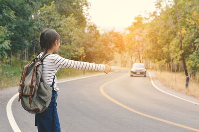 Teenage girl carrying backpack hitchhiking on road amidst trees