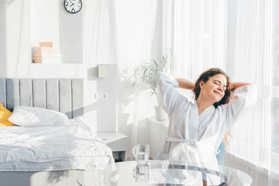 Happy relaxed woman in bathrobe stretches sitting at glass table in spacious bedroom.