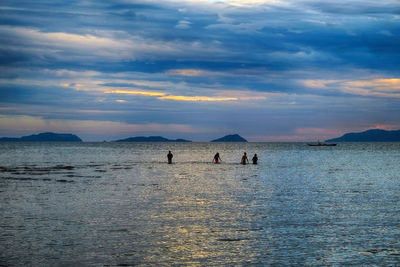 Distant view of people in sea at beach against cloudy sky during sunset