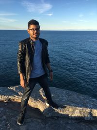 Portrait of young man standing on rock by sea against sky