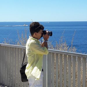 Boy standing on railing by sea against clear sky