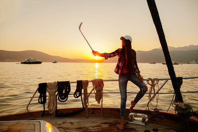 Woman taking selfie with monopod on boat at sea during sunset