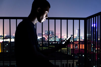Side view of man using mobile phone while sitting in balcony against city at night