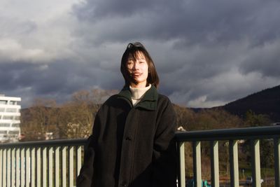 Portrait of smiling young woman standing by railing against sky