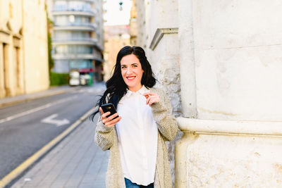 Smiling woman holding mobile phone while standing on footpath in city
