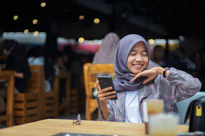 Muslim woman wearing modern hijab smiling and holding smartphone in cafe