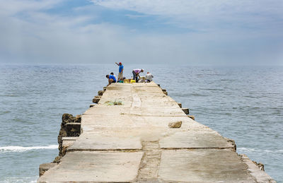 People sitting on groyne by sea against sky