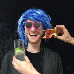 Cropped hand holding drink and pizza slice by man wearing wig and novelty glasses