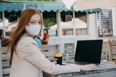 Redhead business woman wearing face mask for virus protection working on the laptop outdoors. young