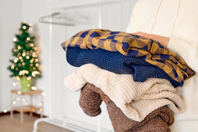 Woman's hands holding stack of warm knitted sweaters in clothes shop with blurred christmas tree. 